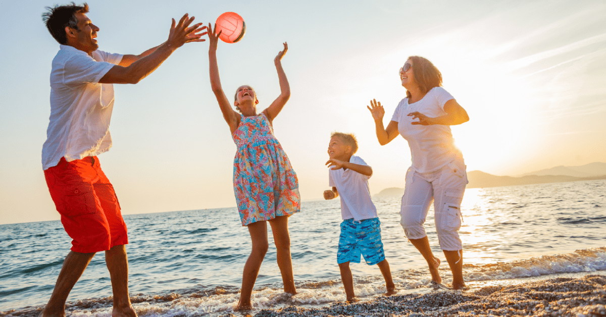 A joyful family of four plays with a ball on the beach at sunset. The scene is lively, with waves splashing around their feet and a warm, golden glow.